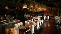 Here's a one-sentence description of the image:A warmly lit dining room at Karawari Lodge features long wooden tables set for a meal, with woven light fixtures and tribal artifacts adding to the rustic ambiance.