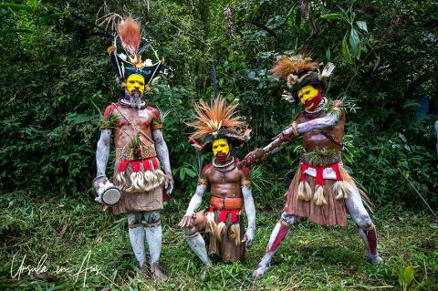 Three colorfully dressed Papua New Guinean tribal members stand in a forest setting, adorned with vibrant face paint, traditional headdresses, and intricate costumes.