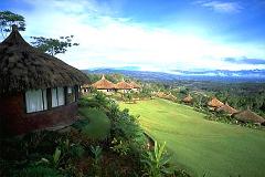 A scenic view of Ambua Lodge, Tari, with thatched-roof bungalows nestled on a lush green hillside overlooking a vast valley and distant mountains under a bright sky.