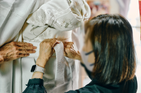 A person is being dressed in a traditional white Japanese wedding kimono, with a masked attendant helping to secure the obi belt.