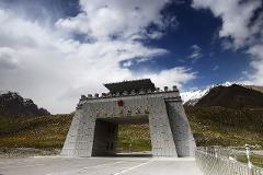 A monumental gate marks the border of the People's Republic of China against a backdrop of mountains and a partly cloudy sky.