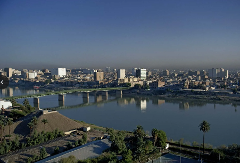 A wide, scenic view of a city skyline with a bridge crossing a large river, framed by palm trees and vegetation in the foreground.