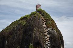 A massive rock formation topped with a building and accessed by a long, winding staircase against a cloudy sky.