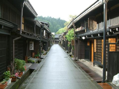 This image depicts a street in Japan with traditional wooden buildings on either side, featuring a wet road surface and a view of a forested area in the distance.