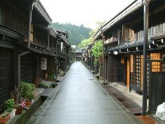 This image depicts a street in Japan with traditional wooden buildings on either side, featuring a wet road surface and a view of a forested area in the distance.