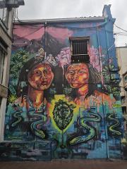 A vibrant mural featuring portraits of indigenous people with floral headpieces, intertwined with snakes and a geometric heart, set against a backdrop of mountains and lush vegetation.