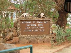 A welcome sign for Tsavo East National Park is adorned with animal skulls, featuring the park's name, area (13,747 sq. km), and altitude (2,050 ft ASL), surrounded by natural elements like rocks and trees.