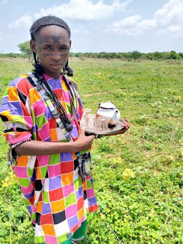 A person in a colorful checkered garment stands in a grassy field holding a tray with a teapot and glasses.