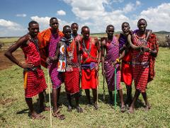 A group of eight Maasai men stand in a grassy field, dressed in traditional red and blue plaid shukas and colorful beaded necklaces, with a blue sky and scattered clouds in the background.