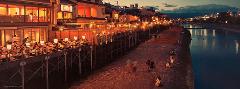 A captivating night scene showcases illuminated restaurants on stilts overlooking a riverbank where people stroll and relax.