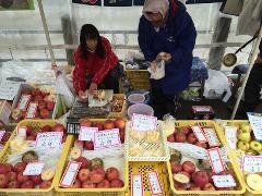 A street vendor sells fresh apples and sliced apples in baskets with Japanese signs and prices. A woman is peeling apples, and another is bagging the sliced fruit.