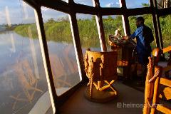 Im Inneren des Kreuzfahrtschiffs Sepik Spirit bedient ein Mann die Steuerung, während er durch große Fenster auf einen Fluss und üppige grüne Vegetation blickt. Aufwendig geschnitzte Holzmöbel verleihen dem Innenraum eine kulturelle Note.