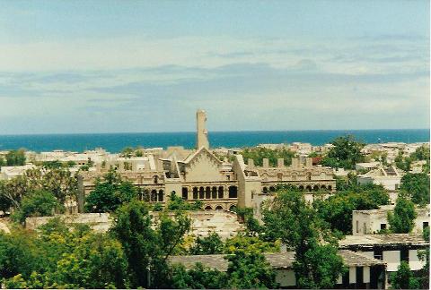 Tur privat de 3 zile în Mogadiscio (istorie, cultură și coastă) An elevated view showcases the weathered, damaged Italian colonial architecture of Mogadishu against a backdrop of the sea and sky, interspersed with lush trees.