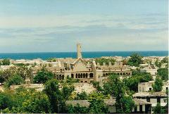 Tur privat de 3 zile în Mogadiscio (istorie, cultură și coastă) An elevated view showcases the weathered, damaged Italian colonial architecture of Mogadishu against a backdrop of the sea and sky, interspersed with lush trees.