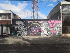 This image shows a mural with three black and white portraits on a wall, with a telecommunication tower in the background and power lines above.