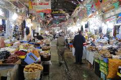 This is a bustling indoor market scene filled with vendors selling various goods, particularly food items such as sweets, nuts, and dried fruits, with numerous shoppers and overhead signage in Arabic.