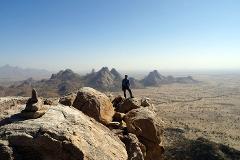 Ein einsamer Wanderer steht auf einem Felsvorsprung und überblickt unter klarem Himmel eine weite, trockene Landschaft mit einzigartigen Felsformationen.