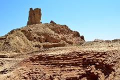A crumbling, ancient structure of layered brick and stone rises against a clear blue sky, showing significant erosion and the passage of time.