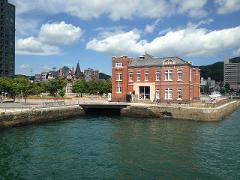 A sunny day view of a red brick building on a stone pier, connected to the mainland by a bridge, with water in the foreground.