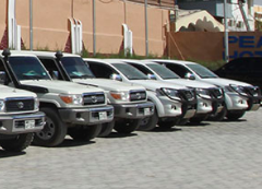 A row of white SUVs, possibly Toyota Land Cruisers, are parked in a lot next to a building labeled "Peace Hotel."