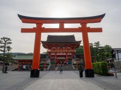 A vibrant orange torii gate stands prominently, framing a traditional Japanese building with intricate details and people exploring the scene.
