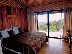 A cozy bedroom with a king-sized bed, wood paneling, and a unique zigzag patterned ceiling, with sliding glass doors leading to a deck overlooking lush greenery.