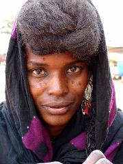 A striking close-up of a woman with dark skin and hair styled in a unique, raised bun, wearing a black headscarf with purple accents and ornate earrings, likely participating in the Gerewol festival.