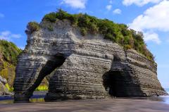 A massive rock formation with layered strata stands majestically on a beach, featuring archways carved by erosion, crowned with vegetation, under a blue sky with scattered clouds.