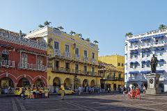 Excursión de un día al Patrimonio Cultural Zenú A vibrant cityscape featuring colorful colonial buildings, a statue, and people in a bustling public square under a clear blue sky.