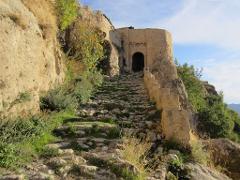 Un chemin sinueux en pierre mène en montée jusqu&#39;à une grande tour en pierre avec une entrée voûtée sombre, flanquée d&#39;un terrain rocheux et de diverses verdures.