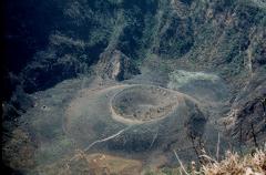 An aerial view captures a volcanic crater nestled amidst lush, green mountains, with a smaller crater inside, surrounded by a barren, ashy landscape.