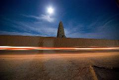 Eine nächtliche Ansicht eines Lehmziegelbaus hinter einer Mauer, mit einem hellen Mond am Himmel und Lichtstreifen vorbeifahrender Fahrzeuge.