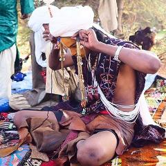 A man dressed in traditional clothing is seated on a rug, adorned with necklaces and jewelry, as he prepares for the Gerewol festival.