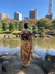 Traditional Japanese Wedding Ceremony & Kimono Experience – 1-Day Tokyo Tour A woman in a colorful kimono stands by a pond in a park, with buildings and the Tokyo Skytree visible in the background.