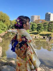 Traditional Japanese Wedding Ceremony & Kimono Experience – 1-Day Tokyo Tour A woman in a traditional Japanese kimono with floral hairpiece stands by a pond in a park, looking towards the buildings in the distance.