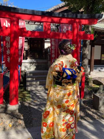 A woman in a traditional Japanese kimono stands before a red torii gate, facing a shrine, with flowers in her hair.