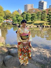 Traditional Japanese Wedding Ceremony & Kimono Experience – 1-Day Tokyo Tour A woman in a beautiful, colorful kimono stands by a pond in a Japanese garden with trees and buildings in the background, possibly part of a traditional wedding ceremony experience.