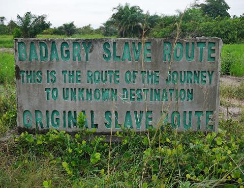 A sign marks the "Badagry Slave Route," stating "This is the route of the journey to unknown destination, original slave route," surrounded by green vegetation against a cloudy sky.