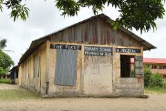 An old, weathered building, identified as "The First Primary School in Nigeria Founded in 1845", stands with a corrugated metal roof and partially boarded windows.
