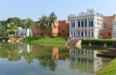 A serene view of Ahsan Manzil in Dhaka, Bangladesh, with its ornate architecture reflected in the calm waters, framed by lush greenery and palm trees under a clear blue sky.