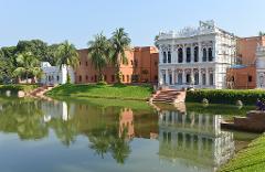 A serene view of Ahsan Manzil in Dhaka, Bangladesh, with its ornate architecture reflected in the calm waters, framed by lush greenery and palm trees under a clear blue sky.