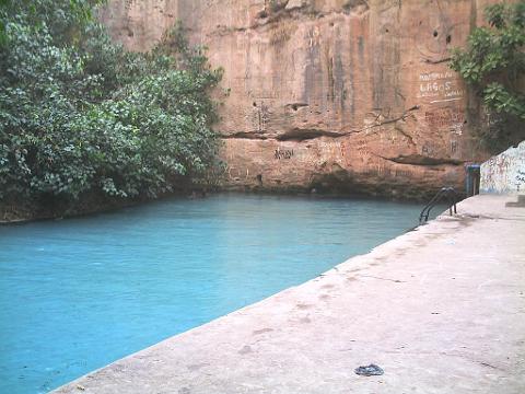 A serene view of a blue-tinted pool bordered by a rocky cliff with graffiti, lush vegetation on the left, and a concrete platform with a ladder on the right.