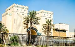 A view of a grand building with palm trees and a black fence, bathed in bright sunlight under a clear blue sky.