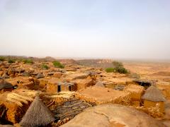 A high-angle view captures a dense village with small, earthy-toned buildings featuring straw roofs and walls, set against a hazy landscape.