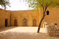A sun-drenched courtyard features a yellow brick building with ornate wooden doors, a leaning tree casting shadows, and a low wall with rounded tops, creating a serene and textured scene.