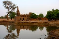 A mud brick mosque is reflected in a pond in front of it, with trees and people nearby.
