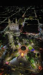 An aerial view showcases a city square illuminated at night, with a grand cathedral and Christmas decorations enhancing the urban landscape.