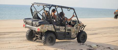 A camouflage-patterned utility vehicle drives along a sandy beach with the ocean in the background.