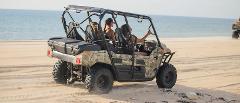 A camouflage-patterned utility vehicle drives along a sandy beach with the ocean in the background.