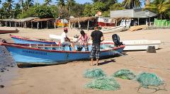 Three men are mending fishing nets next to a blue boat named Maria-Maria on a sandy beach with other boats and buildings in the background.